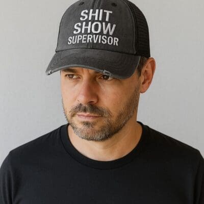 A man with a short beard and mustache wears a black baseball cap that says SHIT SHOW SUPERVISOR in white letters and a plain black t-shirt, standing against a plain light background.