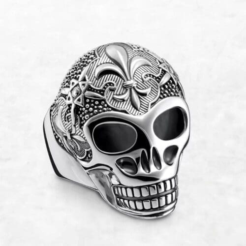 The Blackened Lily Skull Ring features a silver skull with intricate engraved patterns on the top and sides, hollow eye sockets, and detailed teeth, all set against a plain white background.