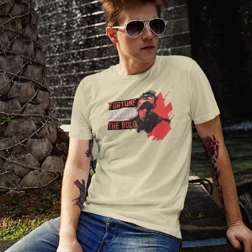 A young man wearing the Fortune Favours The Bold t-shirt and sunglasses sits outdoors near a palm tree