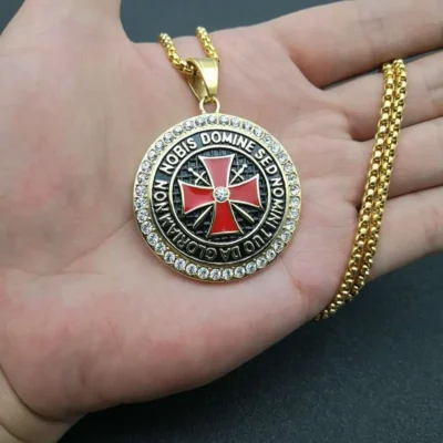 The Crusader Maltese Cross Necklace features a round silver pendant with a diamond-studded edge, a red cross at the center, and Latin text around it, on a silver rope chain. Silver + diamond is written in red at the bottom right.