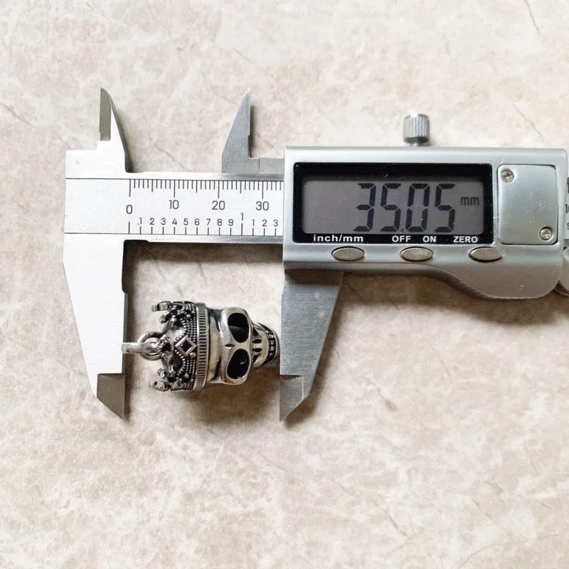 A digital caliper measures the Skull Crown Pendant, showing 35.05 mm on the display. The pendant, designed as a metallic skull with a crown, and the caliper rest on a light-colored textured surface.