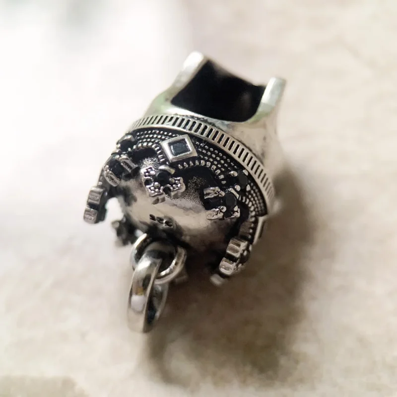A close-up of the Skull Crown Pendant, an ornate, metallic elephant-head design with intricate details and a bottom loop for attaching to chains or bracelets. The background appears blurred.