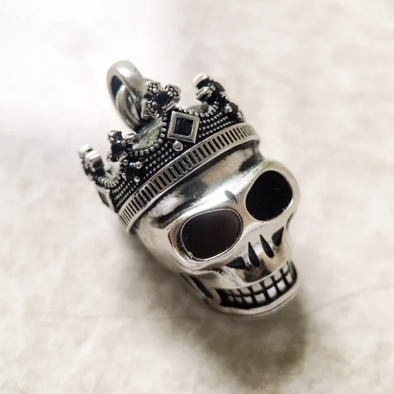 The Skull Crown Pendant is a silver piece featuring a skull with an ornate crown, set against a lightly textured background.