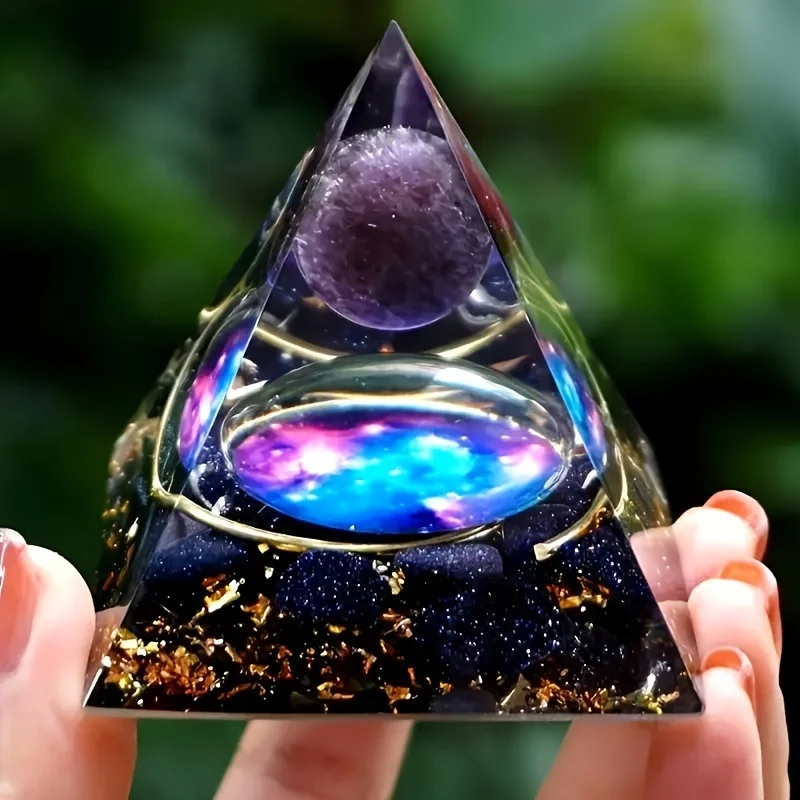 A hand holds the 1pc Galaxy Orgone Pyramid Energy Chakra Reiki Meditation Tool with an Amethyst Ball, blue and pink galaxy design, gold accents, and dark base with gold flakes against a blurred green background.