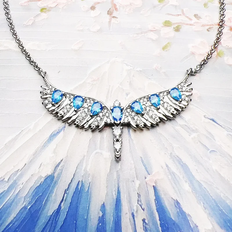 The Phoenix Wing Necklace and Bracelet Jewelry Set features a pendant shaped like outstretched angel wings, adorned with blue gemstones and clear crystals, all hanging symmetrically from a delicate chain.