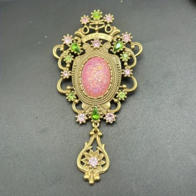 The Baroque Large Vintage Gemstone Brooch features a pink opal-like center, surrounded by pink and green gem accents in ornate gold metalwork, finished with a decorative gold dangle set with a pink gem.