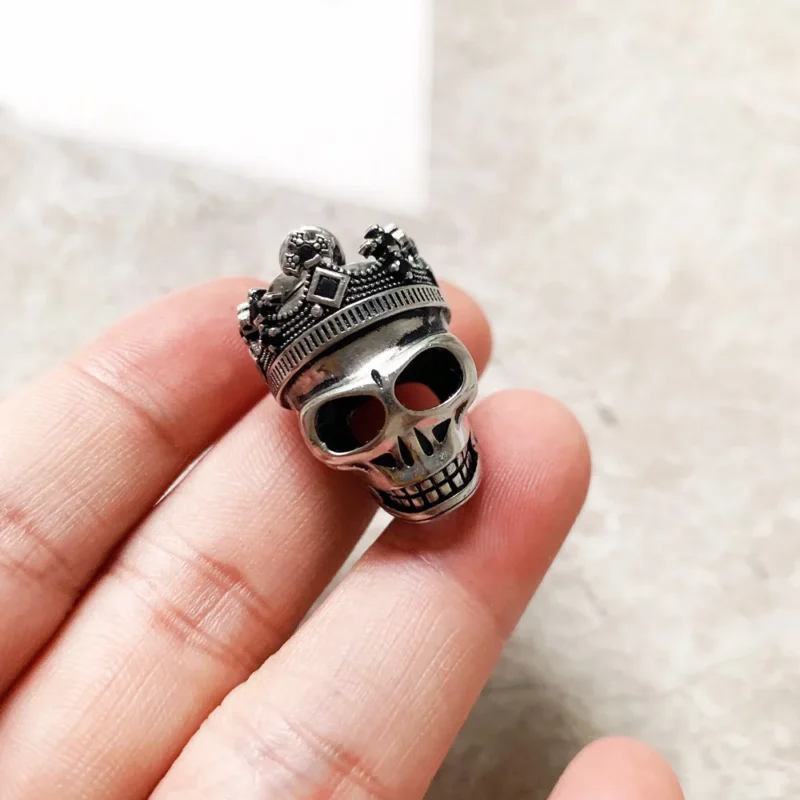 A hand displays the Skull Crown Pendant—a metallic skull with an ornate crown—against a neutral, subtly textured background.