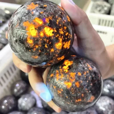 A hand holds two Dragon Stone Yooperlite spheres, their dark gray surfaces with glowing orange flecks—Powerful Chakra Energy Wicca Crystals—in front of similar Healing Spiritual Witchcraft stones in a white bin.