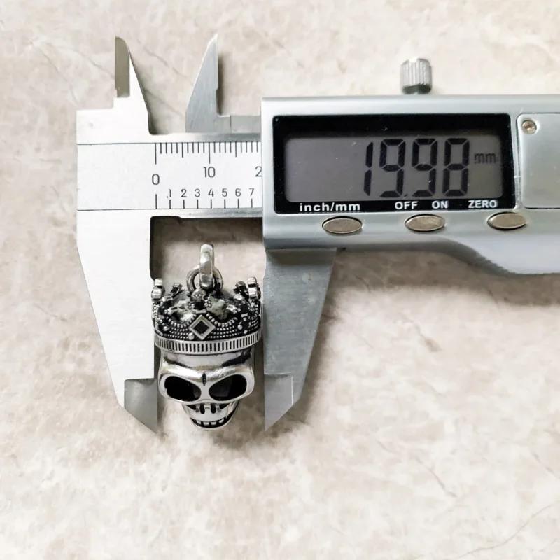 A digital caliper shows the Skull Crown Pendants width as 19.90 mm. The metal pendant is placed on a light, textured surface.