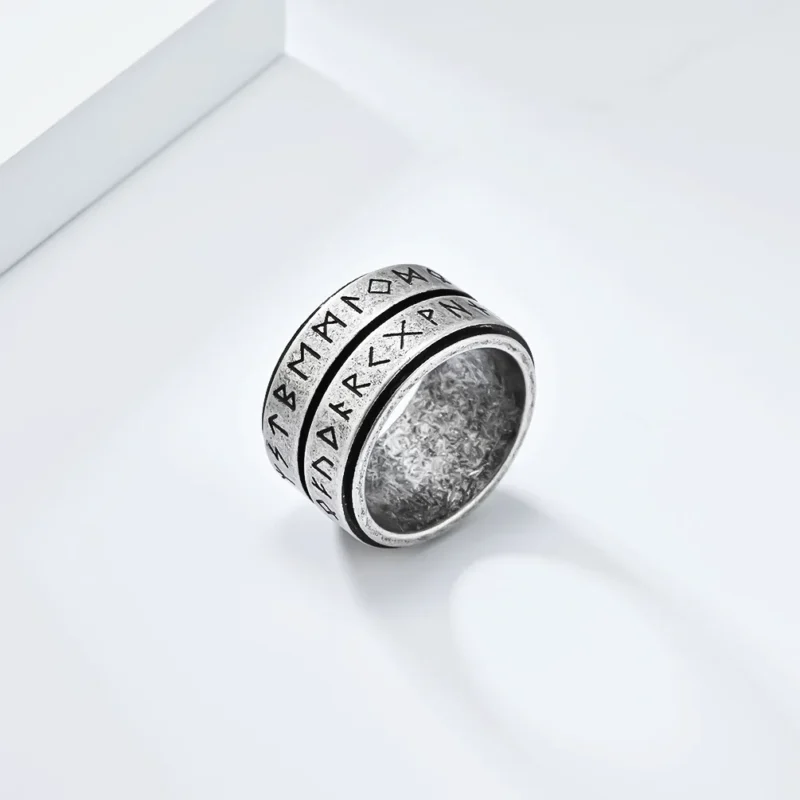 The Nordic Viking Rune Spinner Ring is a wide, silver-colored ring with a worn, textured finish and two rows of engraved runic symbols around its exterior.