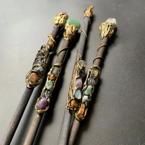The Quartz Point Crystal Wand – Medieval Magic and Mindfulness is displayed on a dark surface, its gold wire and ornate gemstone decorations highlighted by colorful crystals and stones.