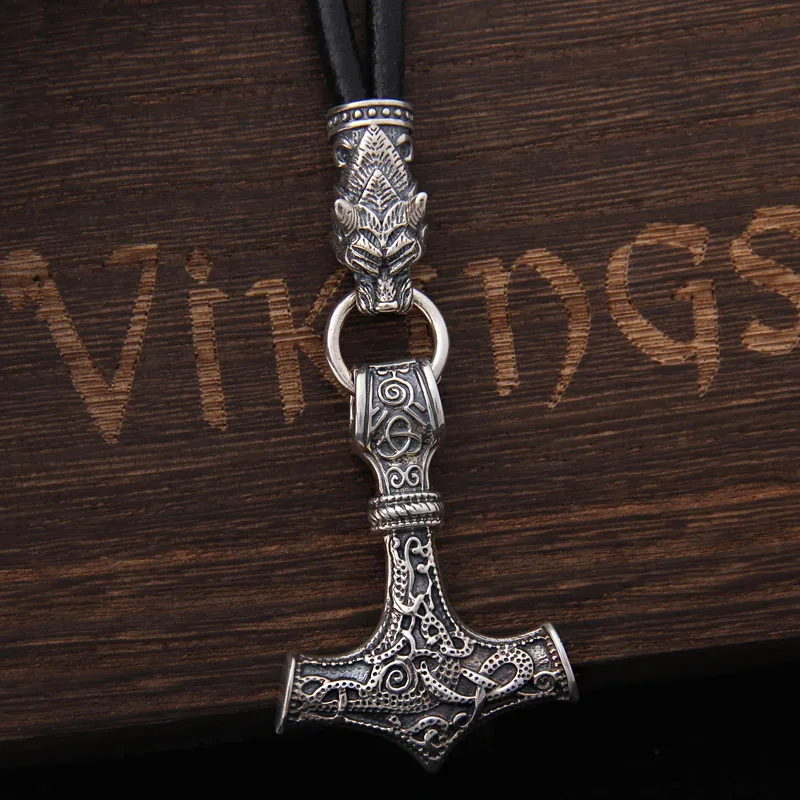 The Viking Thor’s Hammer Pendant with Wolf Head – Norse Jewellery features intricate designs on a silver pendant, suspended from a black cord with a wolf head clasp and displayed on an engraved wooden surface.