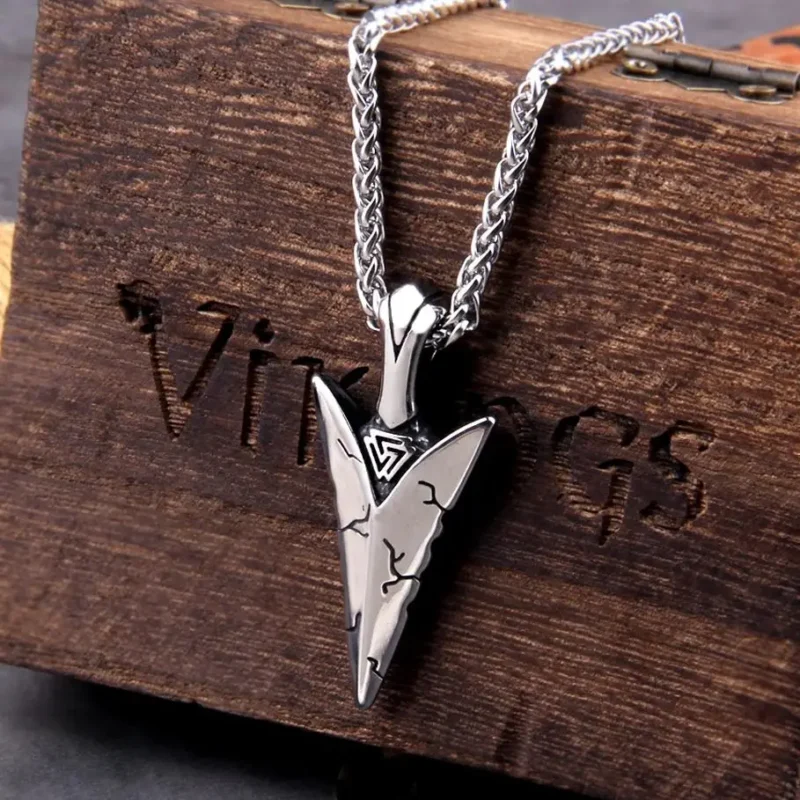 The Viking Spear Necklace features a silver arrowhead pendant with engraved lines and a top triangular symbol, hanging from a braided chain, and is displayed on a wooden box with partially visible engraved text.