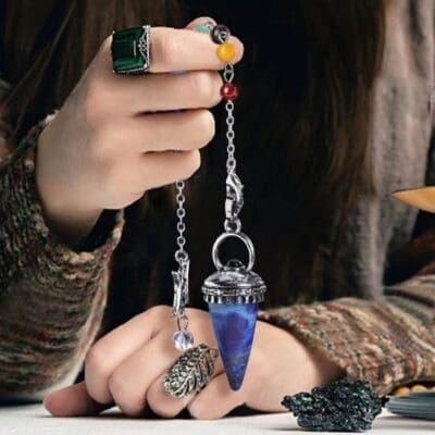 A hand with rings holds a silver chain featuring colourful beads and a blue CSJA Natural Stone Cone Pendulum—Reiki Amethyst Pink Crystal Gem Stone Pendulum for Dowsing, Divination, and Healing Quartz H624. A textured jumper and dark décor blur in the background.